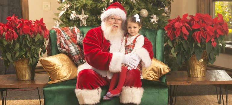 A heartwarming moment of Santa Claus sitting with a young girl on a festive green chair surrounded by poinsettias and a Christmas tree.