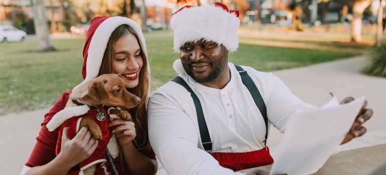 A joyful couple dressed as Santa and Mrs. Claus sharing holiday cheer with their dog in a sunny park setting. Move before Christmas or not, the spirit of the season is infectious.